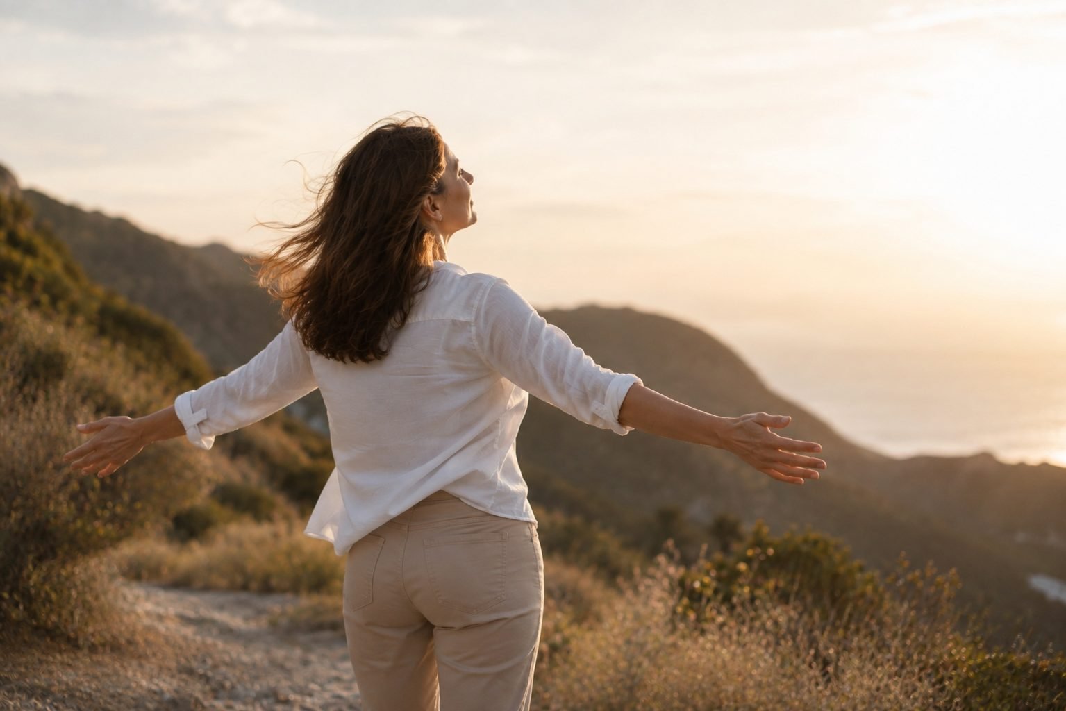 Mulher madura de braços abertos ao ar livre, ao pôr do sol, com expressão de liberdade e bem-estar